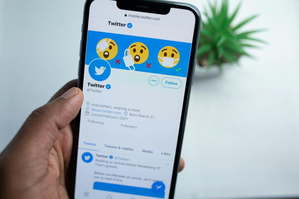 Twitter historia – smartfon pokazujący profil Twittera z emoji w maseczkach, użytkownik trzyma telefon, rozmyte tło z rośliną